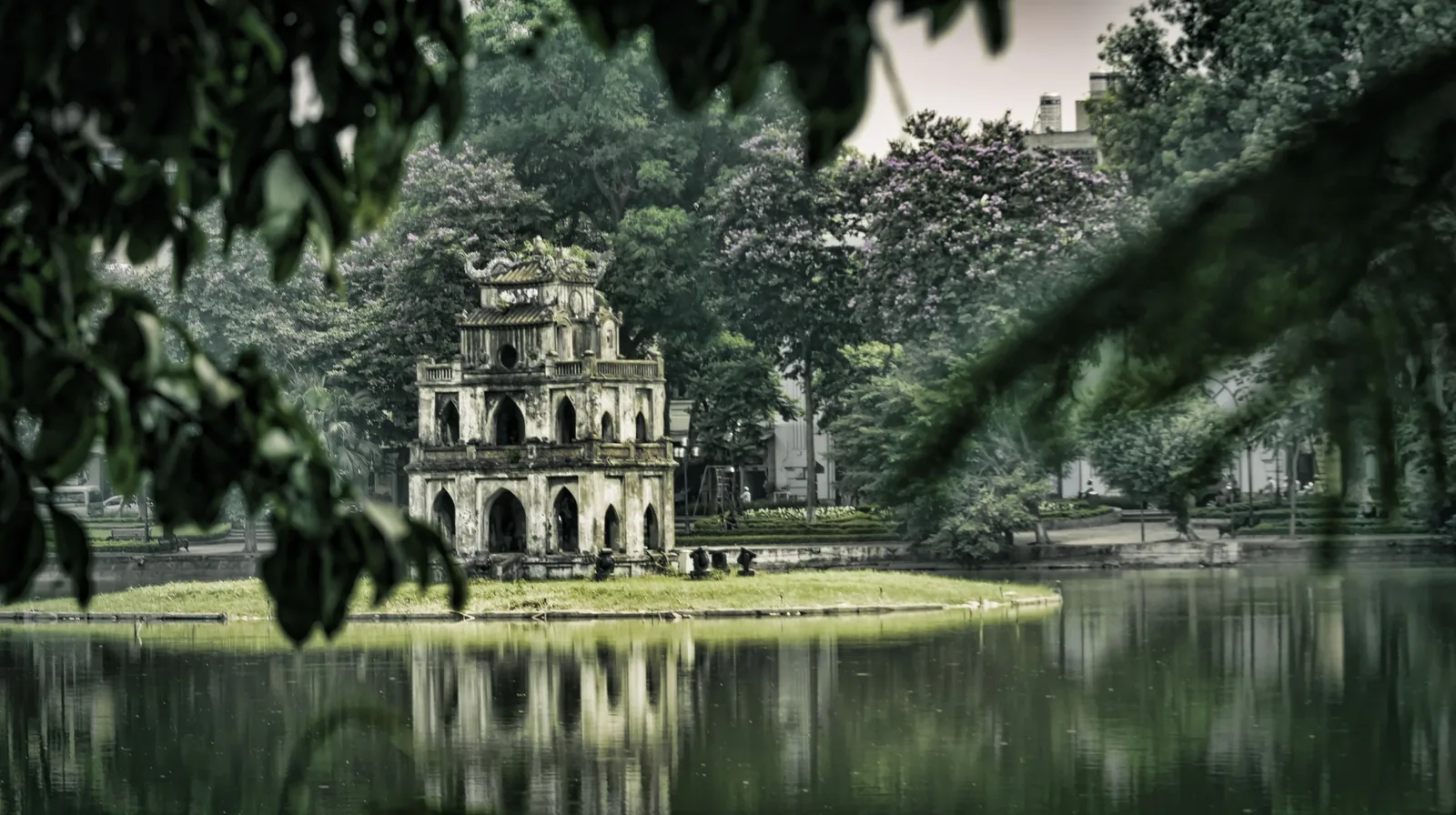 Turtle Tower stands on a small islet in Hoan Kiem Lake, Hanoi, framed by overhanging tree branches.