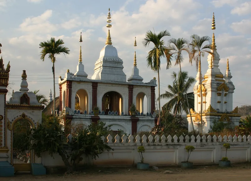 White stupas with tiered spires rise above a walled temple compound with palm trees in Luang Prabang.