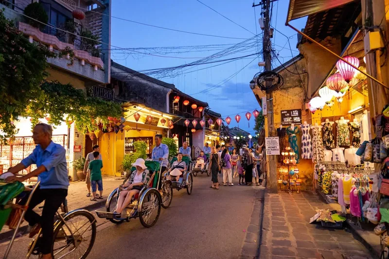 Cyclo drivers pedal tourists past lantern-lit shops in Hoi An's old town at dusk.