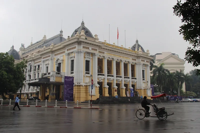 A cyclo rider passes the French colonial facade of the Hanoi Opera House under an overcast sky.