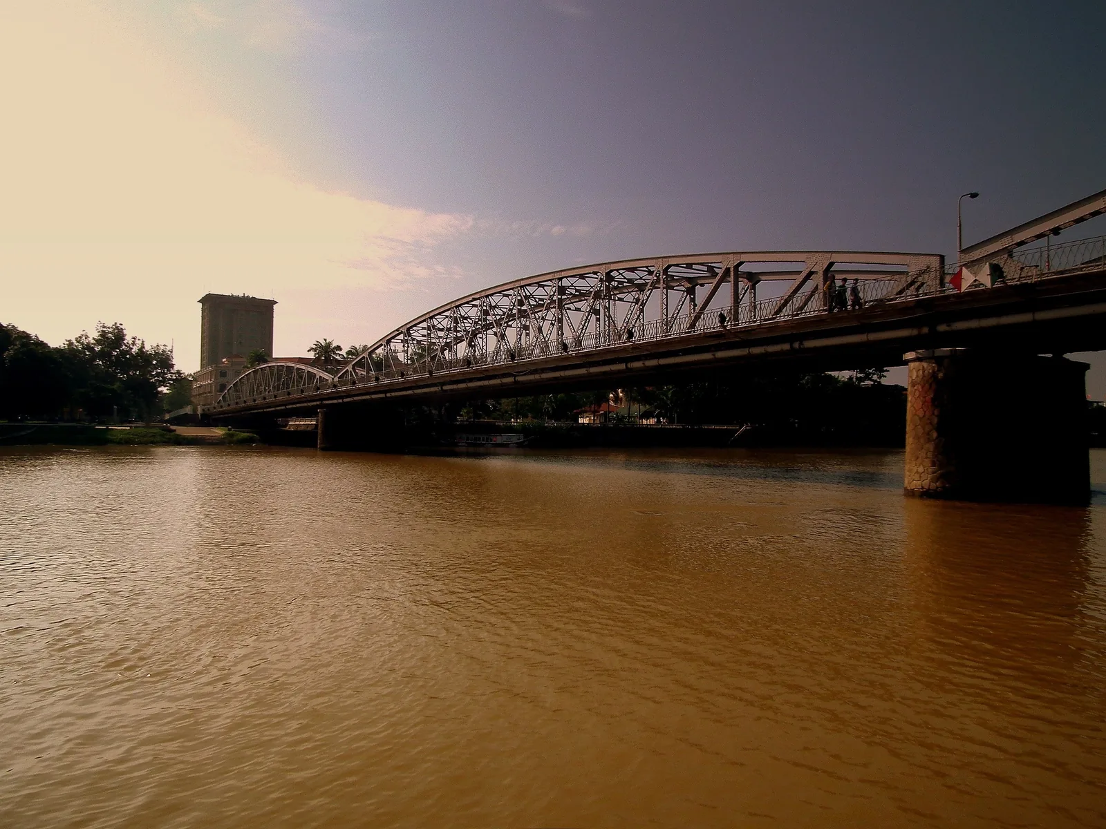 A steel truss bridge spans the muddy Perfume River in Hue under a soft evening sky.