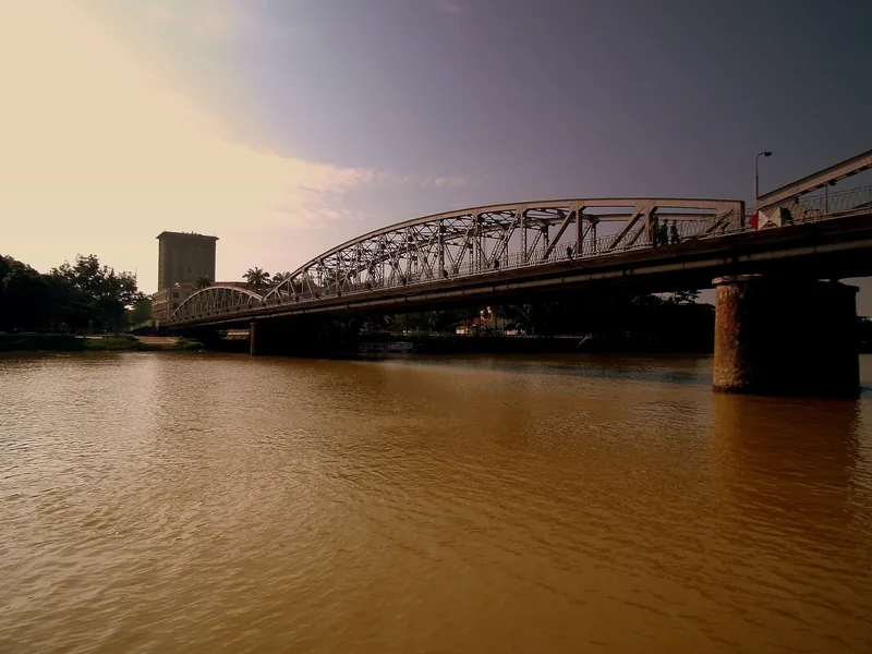 A steel truss bridge spans the muddy Perfume River in Hue under a soft evening sky.
