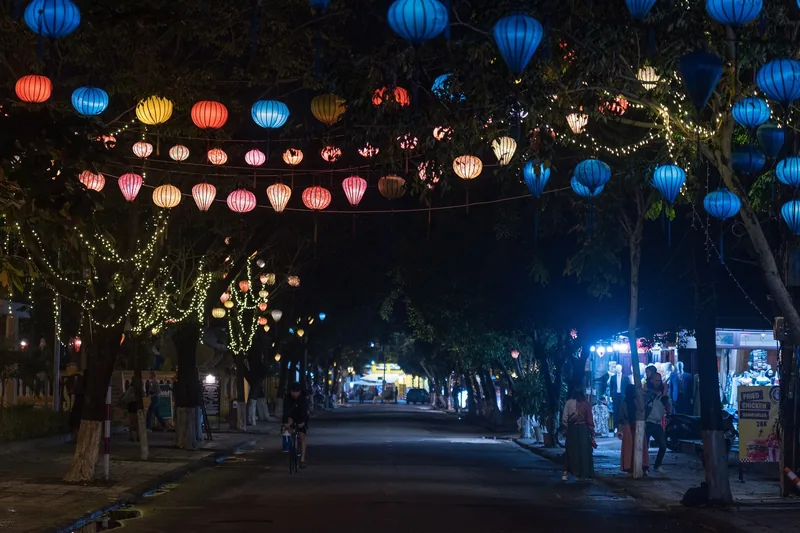 Colorful lanterns strung between trees over a quiet street in Hoi An at night, with a cyclist passing below.