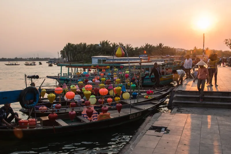 Colorful lanterns decorate boats docked along the Thu Bon River in Hoi An at sunset.