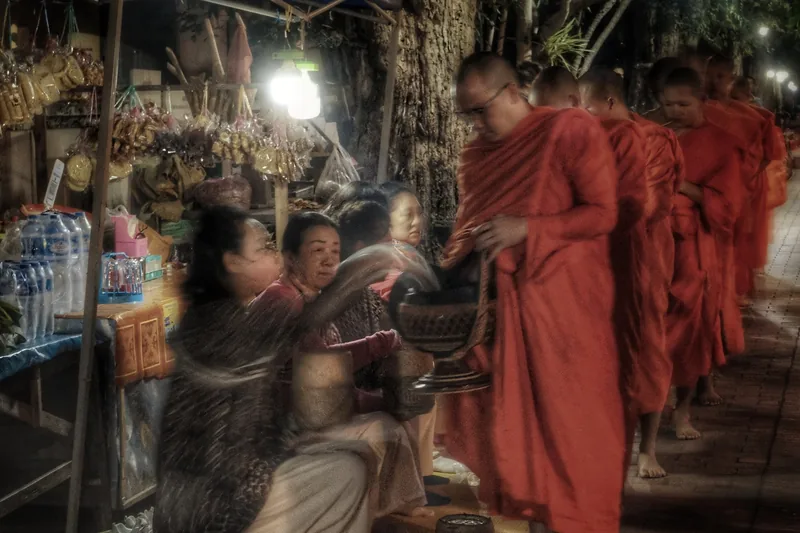 Barefoot monks in orange robes receive alms from kneeling women along a dark street in Luang Prabang.