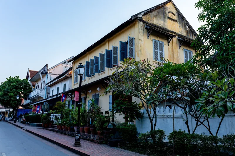 A colonial-era yellow building with blue shutters lines a quiet street in Luang Prabang.