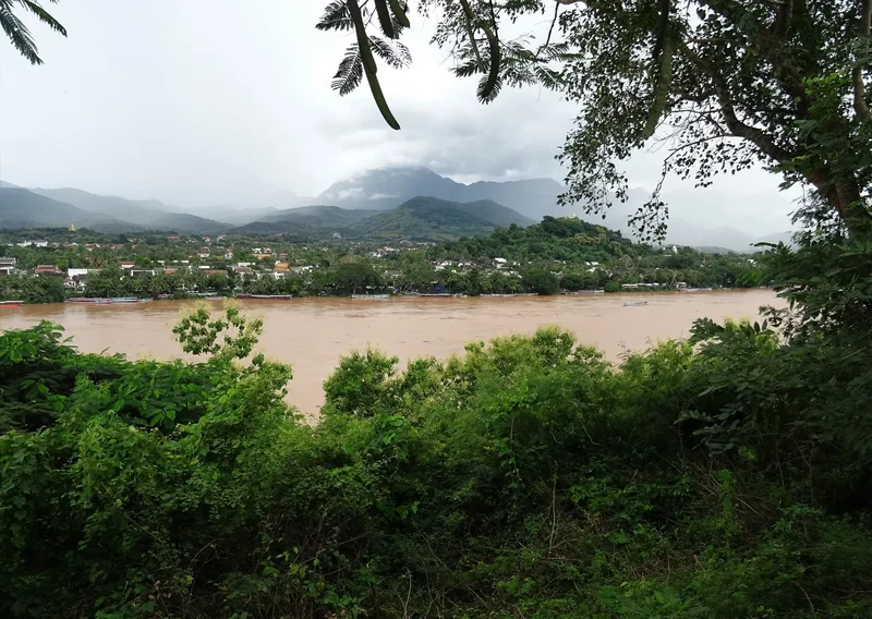 The muddy Mekong River flows past Luang Prabang, seen from a hillside with misty mountains beyond.
