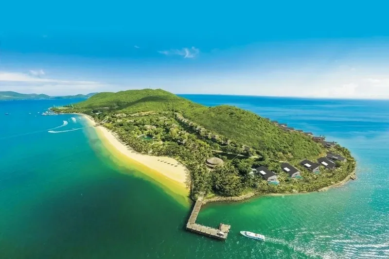 Aerial view of a green forested peninsula with a curved sandy beach and resort villas surrounded by turquoise sea near Nha.
