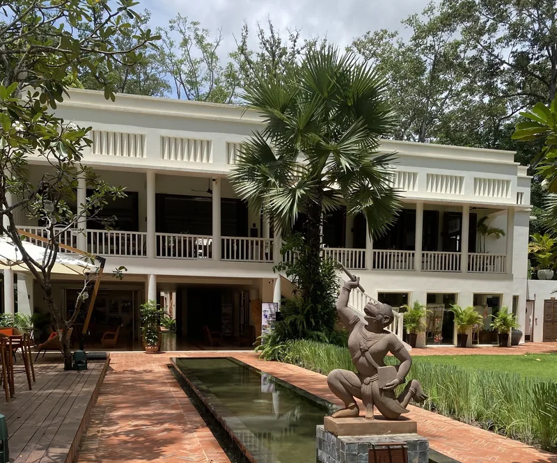 A colonial-style hotel building in Siem Reap features a stone monkey statue beside a reflecting pool and palm trees.
